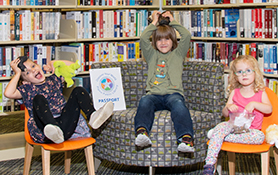 Three kids with a passport in a library setting are ready to Explore Plano Public Library... and discover your world with fun programs and activities all summer long